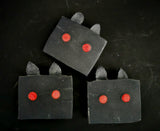 Bars of black soap that have red eyes and black ears meant to look like moth man on a black background.