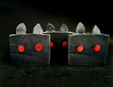 Bars of black soap that have red eyes and black ears meant to look like moth man on a black background.