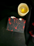 A bar of black soap with red swirls with a dark rose and candle on a black background