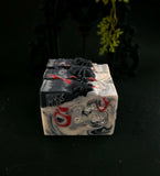 Three stacked handmade soap bars with a marbled black, white, and red pattern, each decorated with a small black spider on top, set against a dark background.