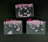 Decorative soap bars in black and purple with flame design and purple flowers topped with a garnet on a dark background