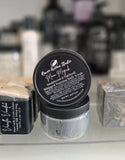 Jar of whipped pampering body scrub with a black lid on a white surface, with other products in the background.