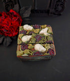 Three handmade marbled soap bars with a black rose motif, including skulls on the top, displayed on a dark surface with a black gothic arch backdrop and a red flower prop.