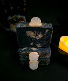 Three stacked rectangular soap bars with a soap crystal ball on top, featuring a dark charcoal and cream marbled pattern set against a dark background with a warm yellow candle glow on the right.