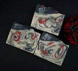 Three handmade soap bars with a gothic spider theme, featuring a swirled white, black, and red design with spiderweb motifs and spider illustrations, displayed on a dark background next to a red spider decoration.