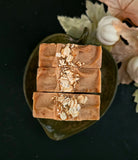 Three rectangular bars of brown soap stacked vertically on a dark green ceramic dish, topped with chopped oats, set against a dark background with decorative autumn leaves.