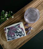 Bar of soap with purple and white design next to a jar of sugar scrub on a wooden tray.