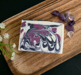 Swirled soap bar with purple and white swirls on a wooden board with amethyst stones and flowers.