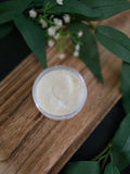 Jar of white sugar scrub on a wooden surface with green leaves and flowers in the background