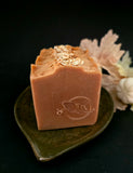 A rectangular bar of brown-colored soap topped with rolled oats, resting on a dark green ceramic leaf-shaped dish against a black background with soft dried flowers.