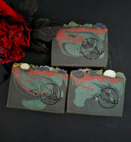 Three handmade marbled soap bars with a black rose motif, including skulls on the top, displayed on a dark surface with a black backdrop and a red flower prop.