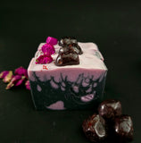 Decorative soap bars in black and purple with flame design and purple flowers topped with a garnet on a dark background