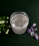 Container of purple sugar scrub with glitter surrounded by purple crystals and white flowers on a dark background