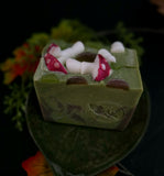 Two rectangular bars of handmade soap with a woodland mushroom theme, featuring green bases with white and red mushrooms, set against a dark background decorated with autumn leaves.