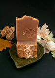 Stacked bars of handmade brown soap with oat flakes, displayed on a dark green ceramic leaf-shaped dish against a black background with autumn leaves and pumpkins.