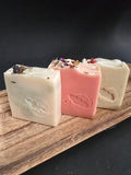 Three soap bars of different colors and textures topped with herbs on a wooden surface with a dark background