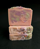 Three stacked handmade soap bars with a dusty pink base, purple and green marbled swirls, and dried botanical inclusions, set against a solid black background.