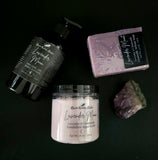 Lavender-themed products including a bottle of lotion, jar of sugar scrub, soap bar, and crystal on a dark background.