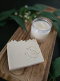 Jar of white sugar scrub and bar of soap on a wooden board with greenery in the background