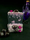 Stack of decorative soap bars in black and purple with flame design and purple flowers topped with a garnet on a dark background