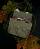 Two rectangular bars of handmade soap with a woodland mushroom theme, featuring green bases with white and red mushrooms, set against a dark background decorated with autumn leaves.