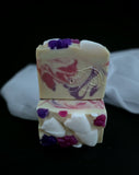 Stacked bars of handmade soap with a cream base, featuring swirls of pink and purple, embedded white ghosts and purple and pink bows on top, set against a black background with a white fabric prop.