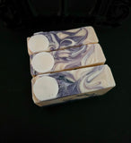 Three rectangular bars of handmade soap with a purple and cream swirl pattern and a full moon motif  arranged vertically on a dark background.