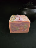 Pink soap with green and purple  swirls with herbs on top on a black background.