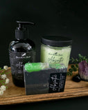 Bottle of lotion, sugar scrub, and bar of soap labeled 'Absinthe' on a wooden tray with a dark background.