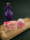 Purple bottle, pink sugar scrub in a clear container, and soap with swril design on a wooden surface