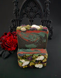 Three handmade marbled soap bars with a black rose motif, including skulls on the top, displayed on a dark surface with a black gothic arch backdrop and a red flower prop.