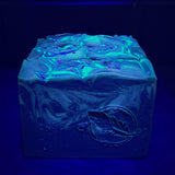 Soap bar in dark purple and silver accents on a black background shown under blacklight. 