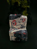 Three stacked handmade soap bars with a marbled black, white, and red pattern, each decorated with a small black spider on top, set against a dark background.