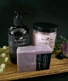 Lavender-themed skincare products including a bottle of lotion, jar of sugar scrub, and soap on a wooden surface with a dark background.