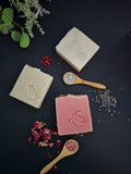 Three bars of tallow soaps with a logo on a dark surface with dried flowers and herbs.