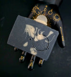 A rectangular black soap bar with gold marbled swirls and a circular embossed design, held in a black hand-shaped prop decorated with gold celestial symbols.