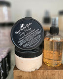 Jar of foaming scrub with a black lid and a bottle of vanilla oil on a wooden surface.