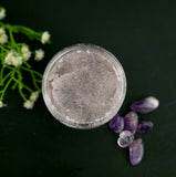 Top-down view of a container of purple sugar scrub with glitter surrounded by purple crystals and white flowers on a dark background