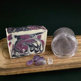 Marbled soap bar with purple and white swirls on a wooden board with crystals and a clear container. of purple sugar scrub.
