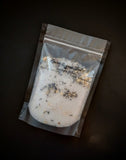 White bath salts with lavender and jasmine buds pictured in a black bag